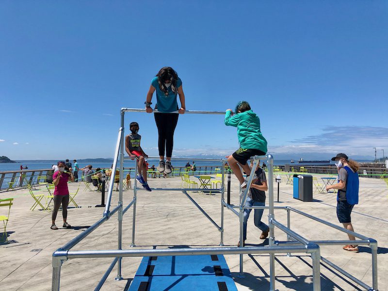 Kids practice parkour on a set of parallel bars while an instructor looks on: one girl with light brown skin looks down while cresting over the top bar. A bluebird sky and the Salish Sea are visible behind them.