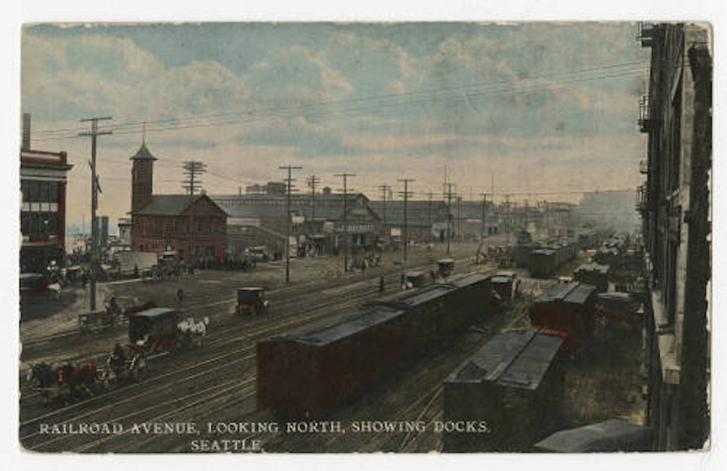 In the foreground are railroad tracks, in the distance are buildings and beyond them the waterfront, the sky is painted blue with some fluffy clouds.