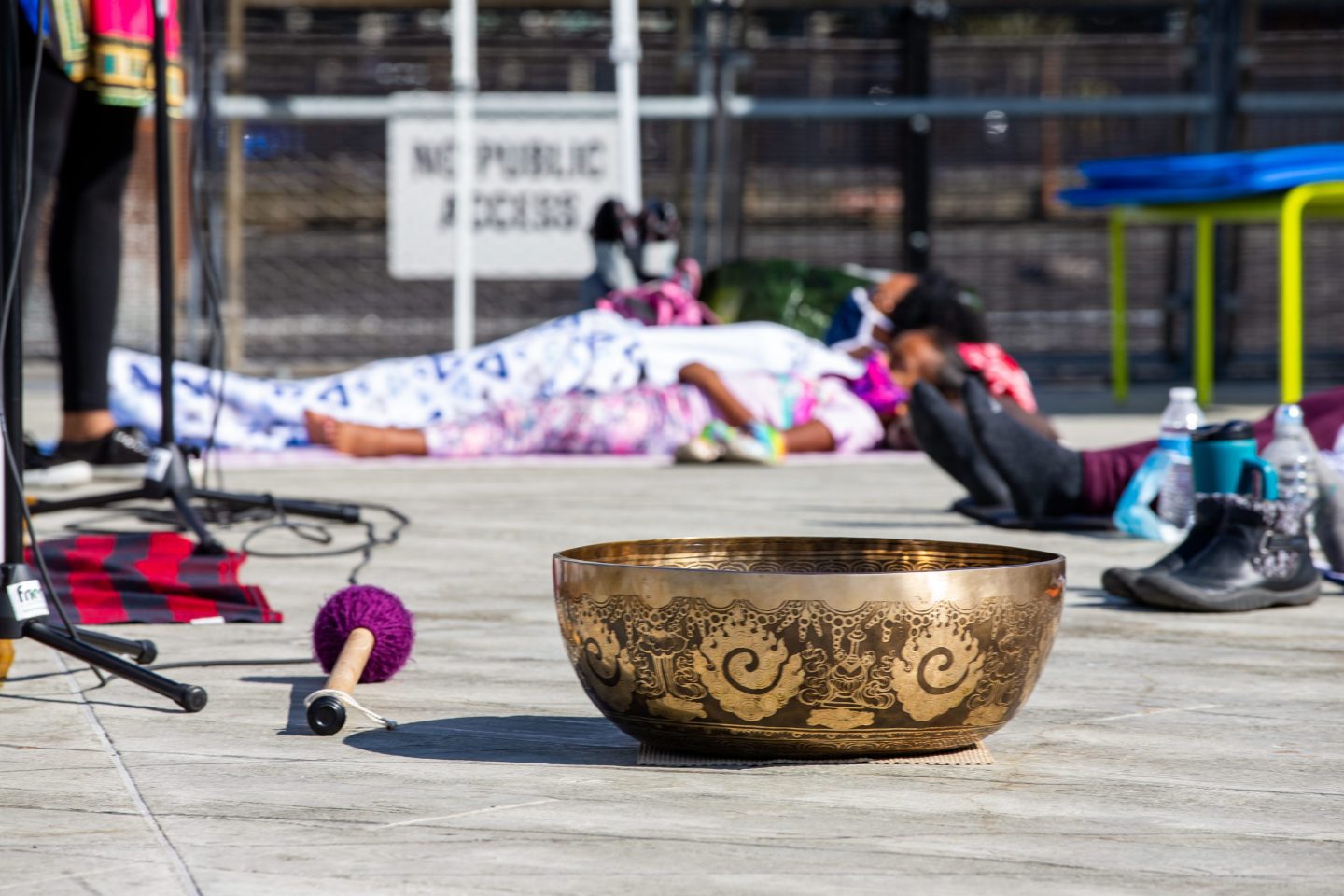 An engraved golden bowl sits on a boardwalk, meditation attendees are visible but blurry in the background, they are lying on the ground.