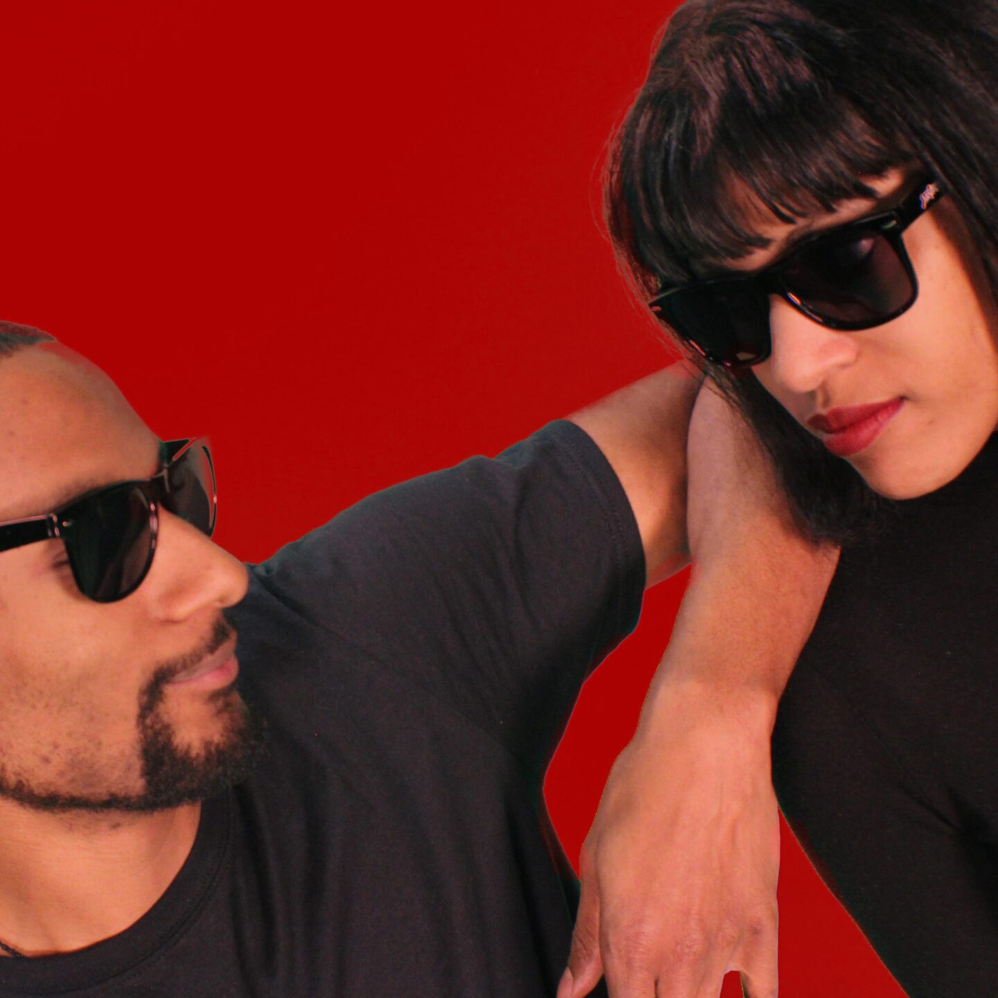 Two cool people, a man and a woman, with black sunglasses and black shirts are looking and leaning at each other in a diagonal fashion vs the framing, the background is vivid red.