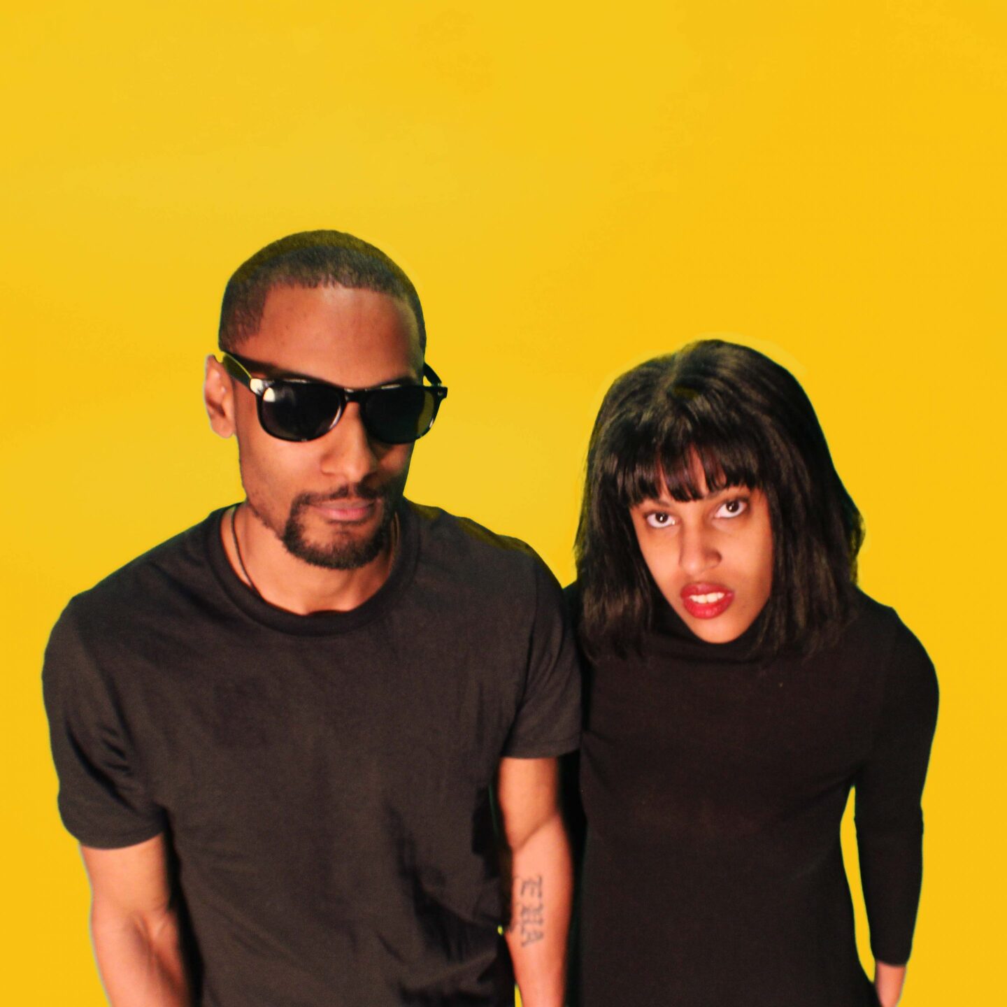 Two cool people, a man and a woman, wearing black shirts are standing next to each other looking up at the camera, the photo framing is cut off at their waists and the background is a vivid yellow.