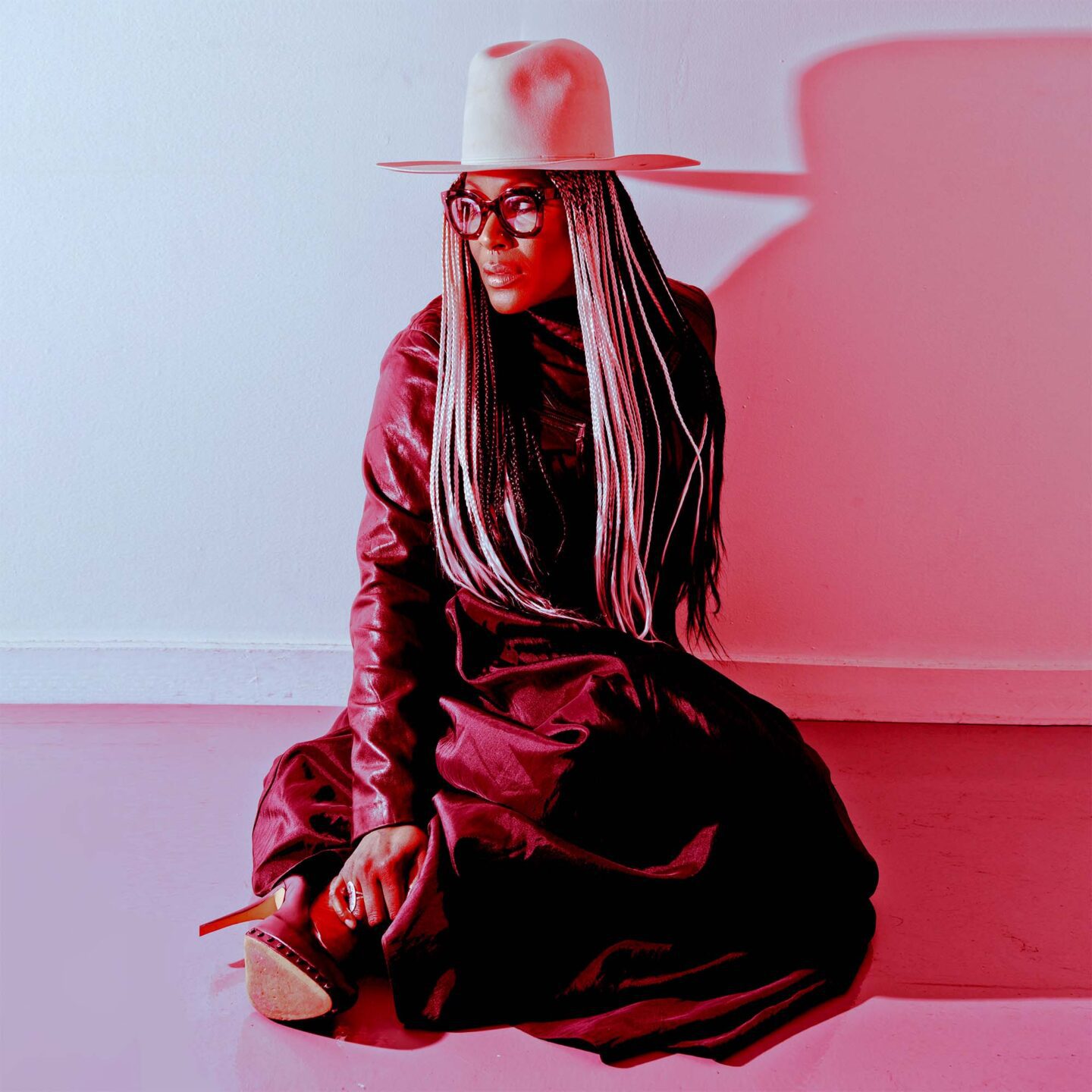 A woman sits with her legs folded to one side on the floor looking off in the distance. She has a dark dress on, a white hat with a brim, and high-heeled shoes. The photo is awash in red light.