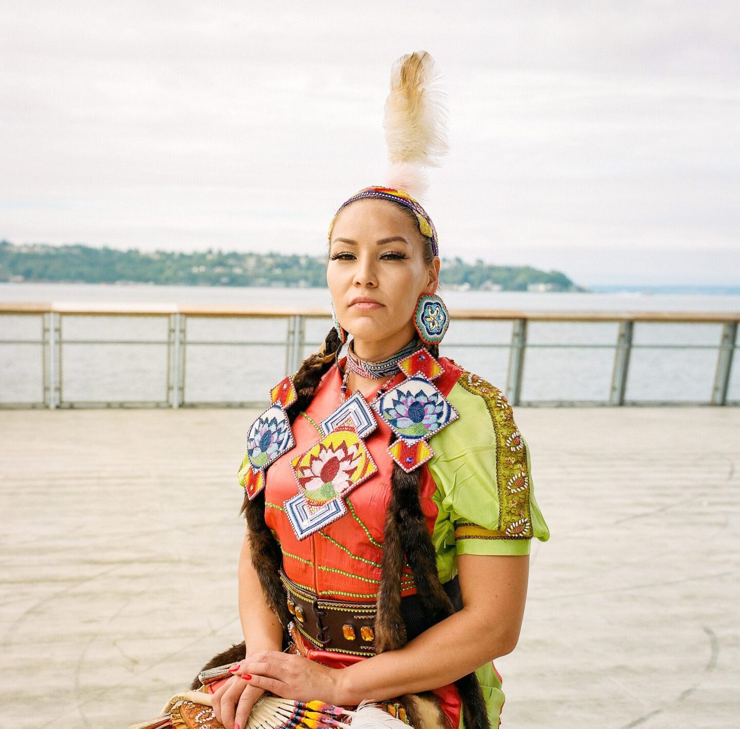 Abriel Johnny -A native woman (Cowichan and Tlingit) stands proudly in a colorful dress of orange and yellow-green, she holds a fan of feathers and stands on the boardwalk of Pier 62. The Salish Sea is visible in the background against a bright cloudy sky.