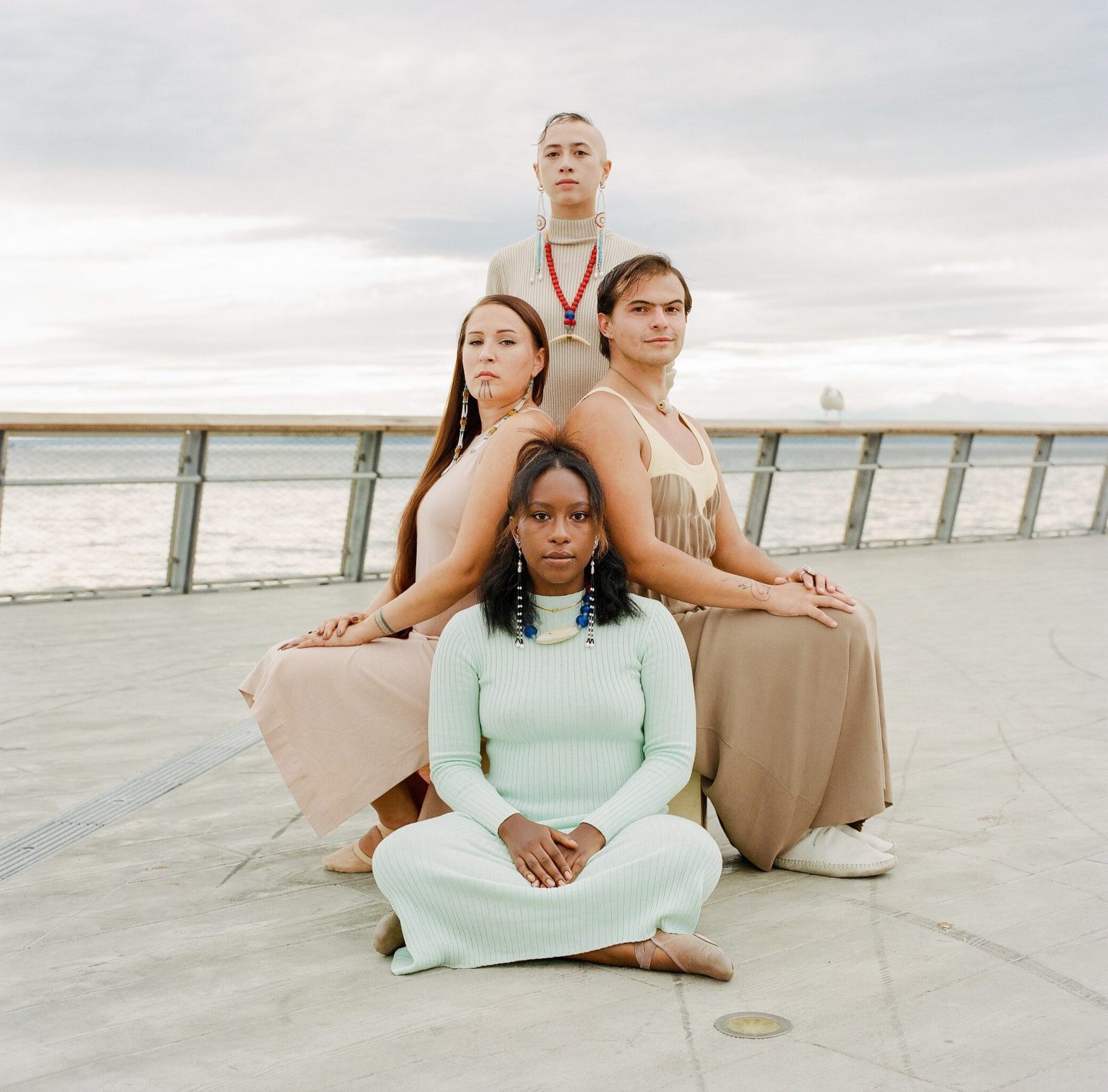 Four native people of a range of skin tones wear pastel colors, sitting and standing on the deck of a pier. The Salish Sea is visible in the background against a bright cloudy sky.
