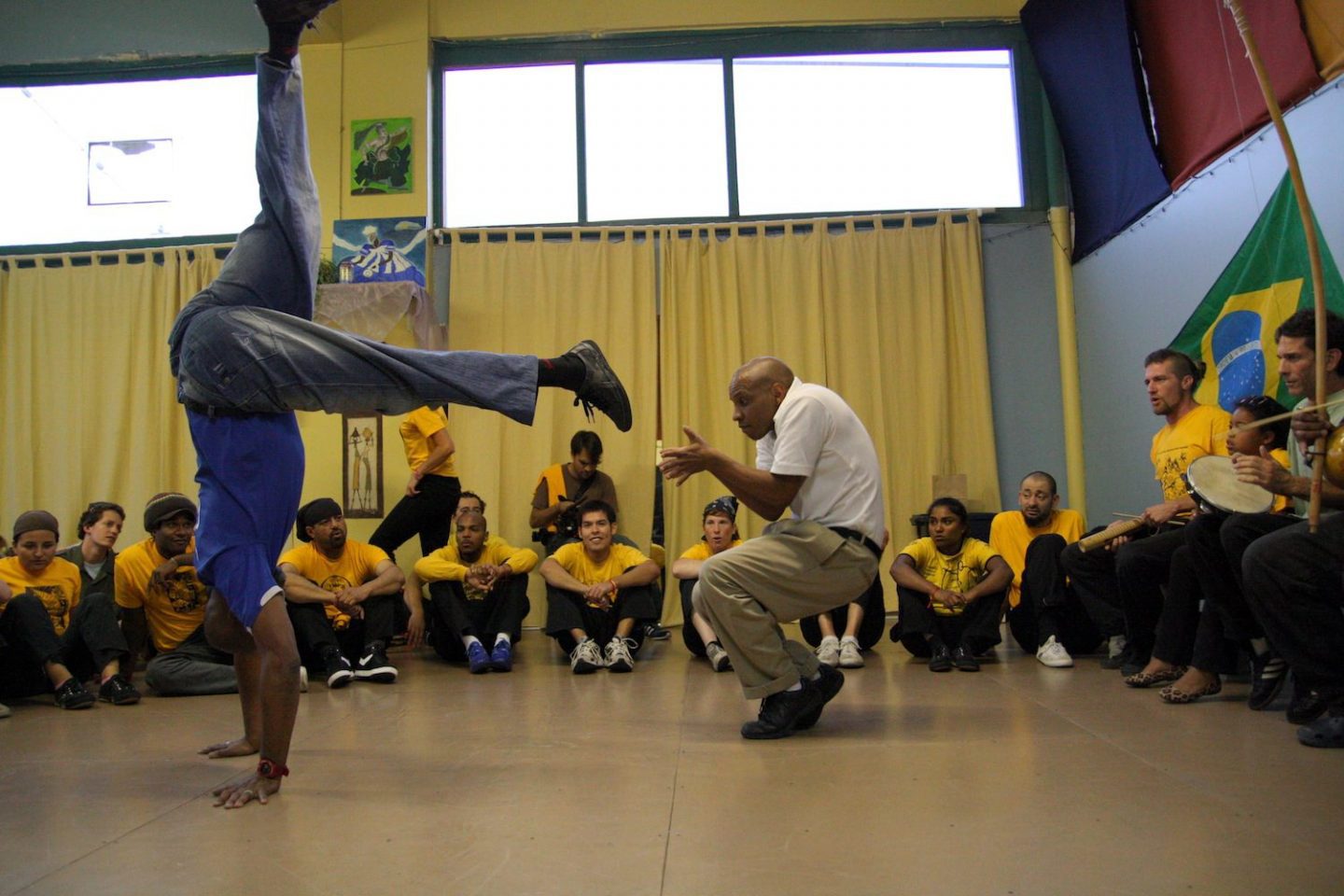 An awesome black man in a crisp white short and khakis crouches while dodging his fellow sparing partner's foot, the other man has two hands on the floor, one leg up high and one leg mid-swing across. Onlookers in yellow shirts and black pants sit in a circle (a capoeira roda) on the floor.