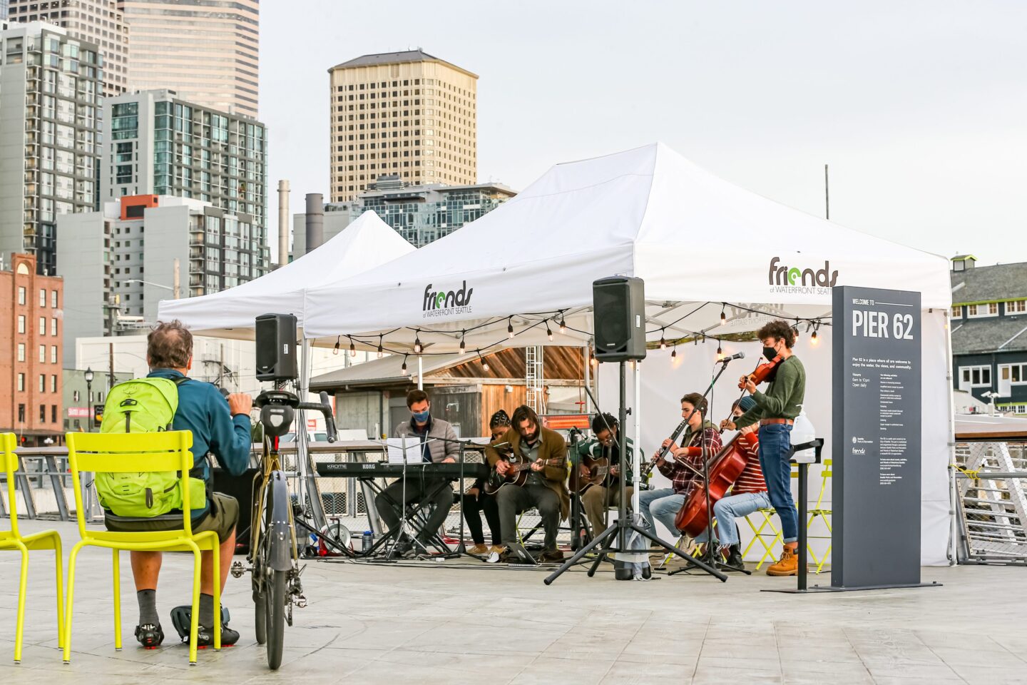 Young-looking musicians sit and stand in a row while a man with a bicycle looks on.