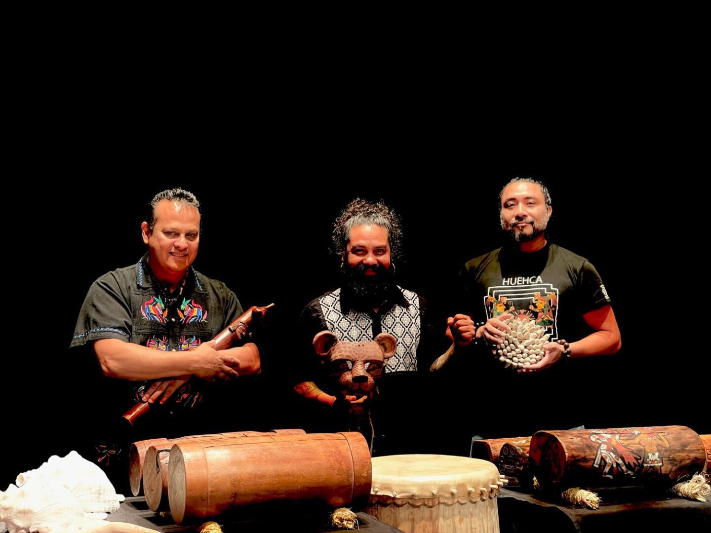 Three smiling middle-aged men with black hair stand in front of a black background, they hold traditional instruments. There is a table with more heritage items on it visible in front of them.