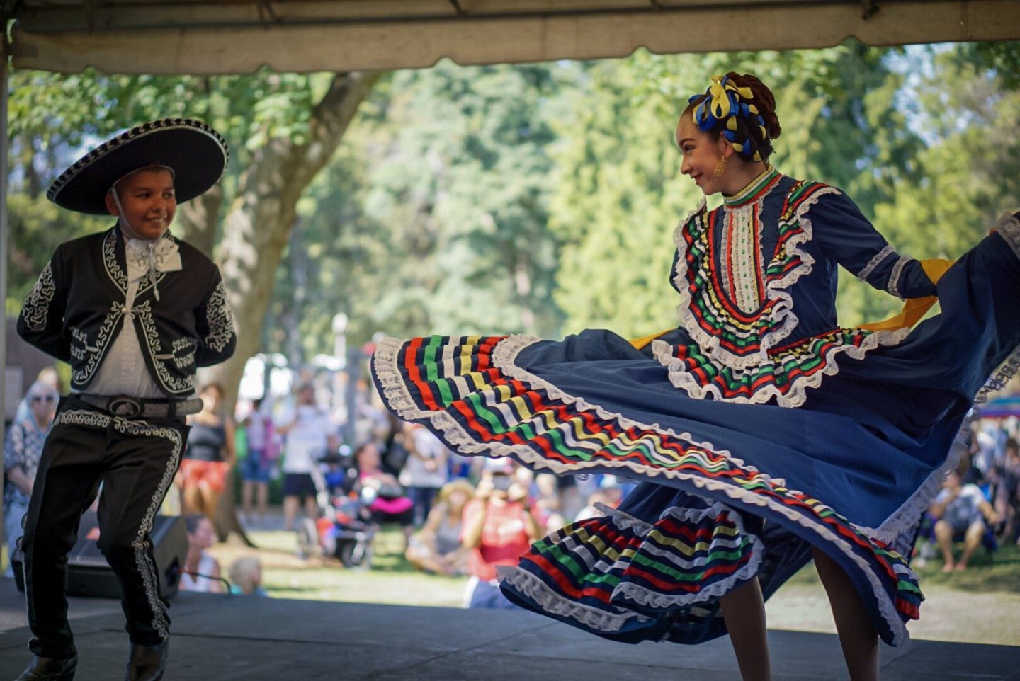 Two adolescents on a stage in traditional Mexican garb, are smiling and looking at each other mid-dance.