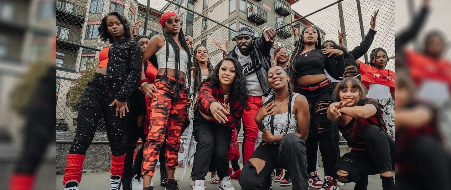KUTTNUT - about a dozen young-looking black women and one man stand or squat while smiling at the camera. They are dressed stylishly in red, black, and white and look ready to dance.