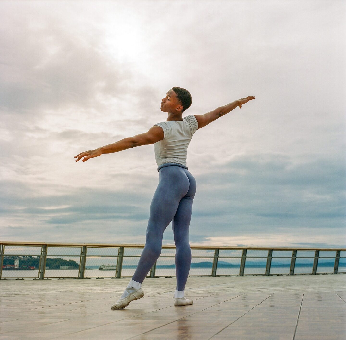 A brown person with short hair, white t-shirt and tight jeans sits and poses on the boardwalk of Pier 62. The Salish Sea is visible in the background against a bright cloudy sky.