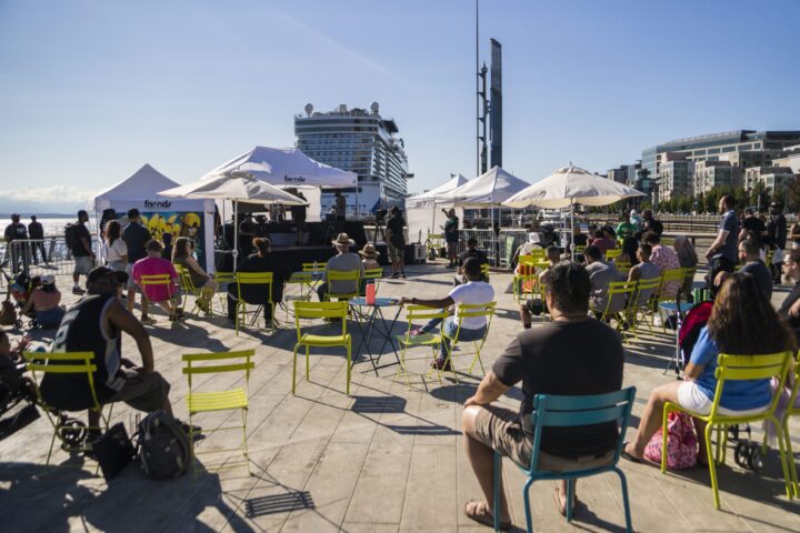 206 Zulu Beat Masters: a diverse audience sits outside on nice patio chairs watching  music makers under a tent. A cruise ship and blue sky is visible in the background with bright sunshine. Photo by Tosin Arasi.