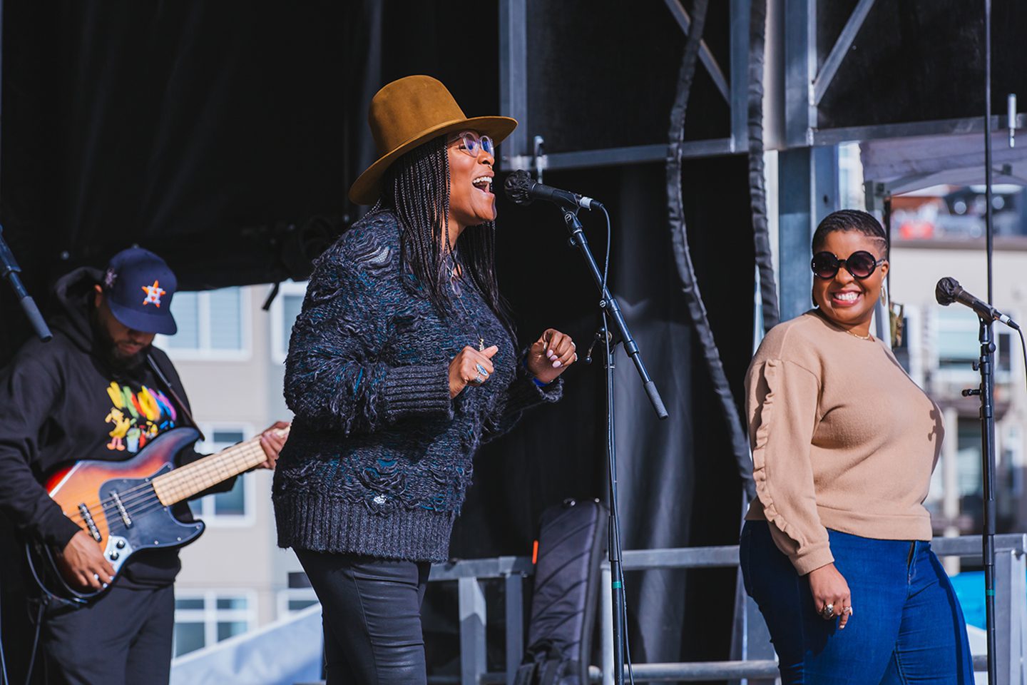 A woman with a light brown hat and complexion sings and smiles into a microphone, she is wearing a black sweater and dark pants. A backup singer looks on smiling in the background along with a guitarist. They are all on a stage (not visible in the picture).