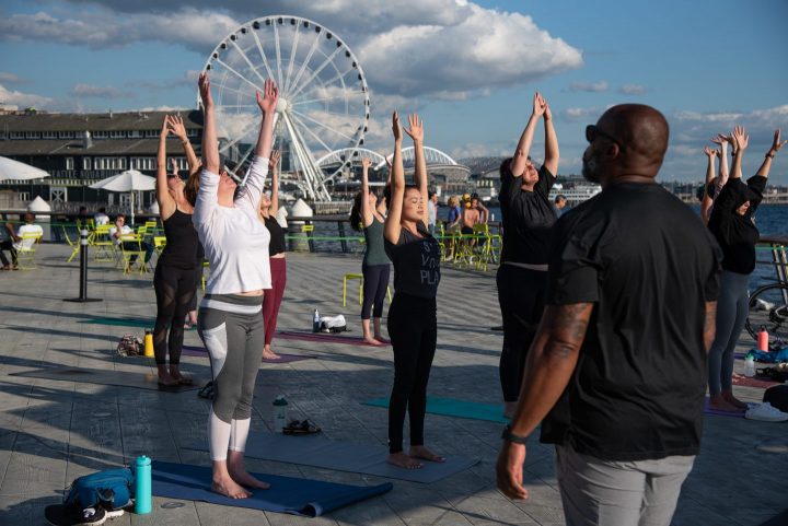 Several students in yoga pose on a pier with the Seattle great Ferris wheel in the background.