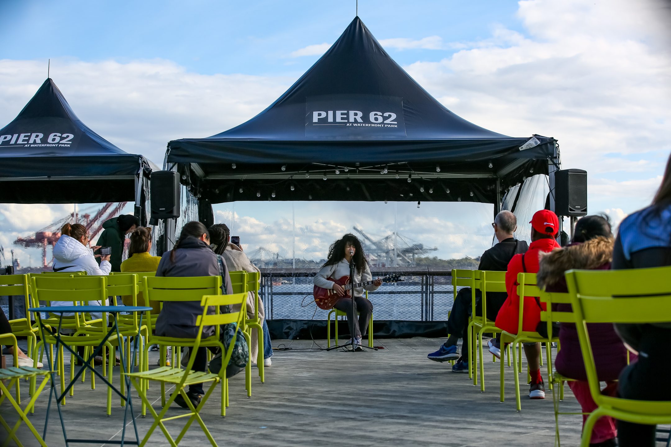 A light caramel-skinned women sits with a guitar, singing into a mic under a fancy black tent. In the foreground are attendees on neon yellow chairs, in the background is the Seattle skyline, Elliott Bay, and fluffy clouds with patches of blue sky.