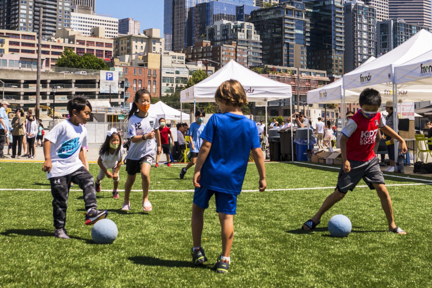 Children play in the sunshine on a soccer field, kicking a ball around.