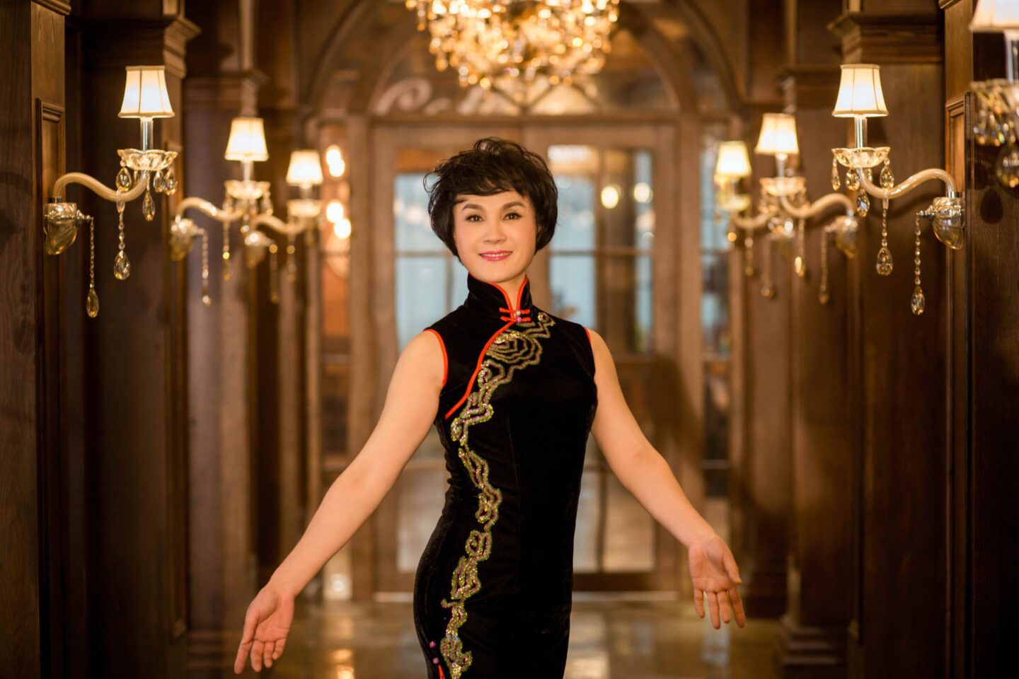Melody Institute dances at Spotlight July 13, 2023. A woman of asian decent wearing a black qipao/cheongsam with gold and red embroidery stands with open arms in front of a hallway lit with soft golden light.