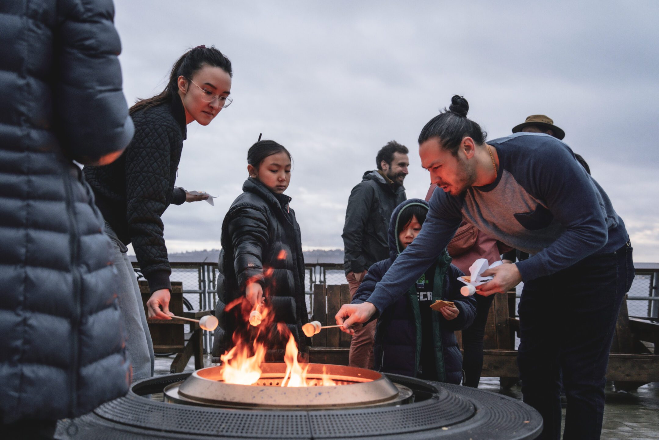 A couple of people lean over a fire pit on Pier 62 to roast marshmallows.