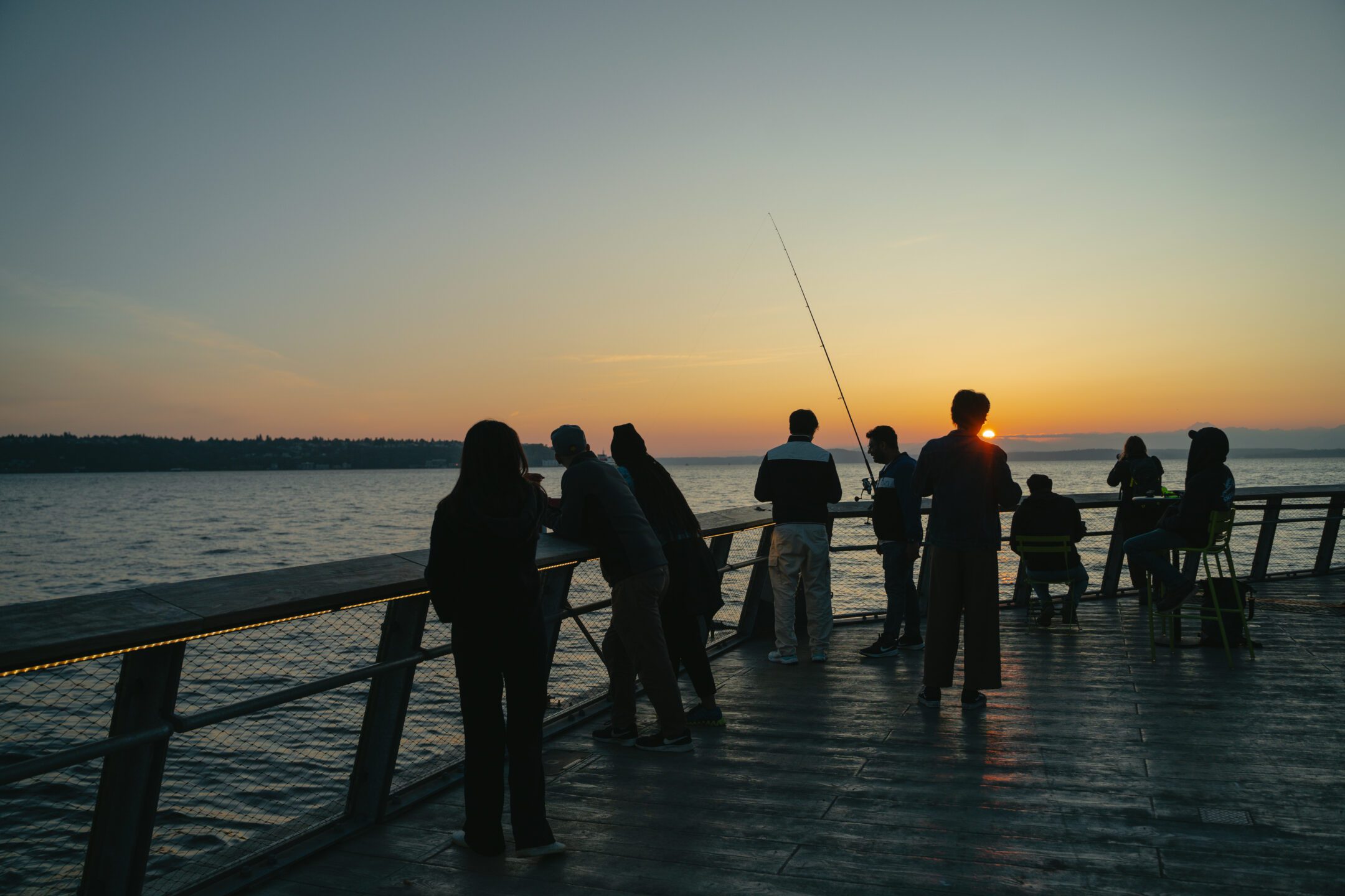 Multiple people leaning over railing at Pier 62. One figure is carrying a fishing rod. The sun is setting over the horizon in the background.