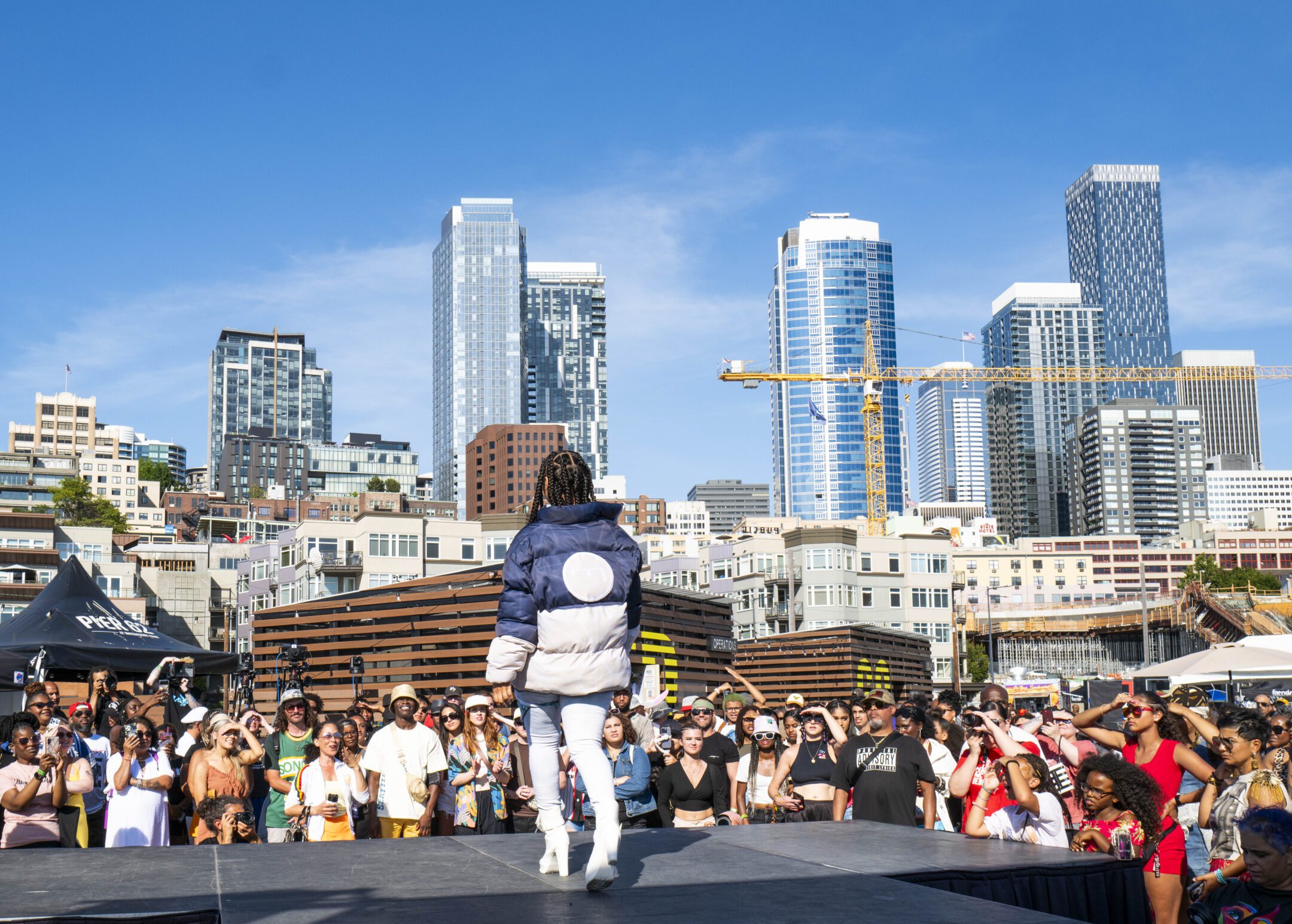 A woman in a blue and white coat and white patnts stands on stage in front of a crowd with the Seattle skyline behind her.