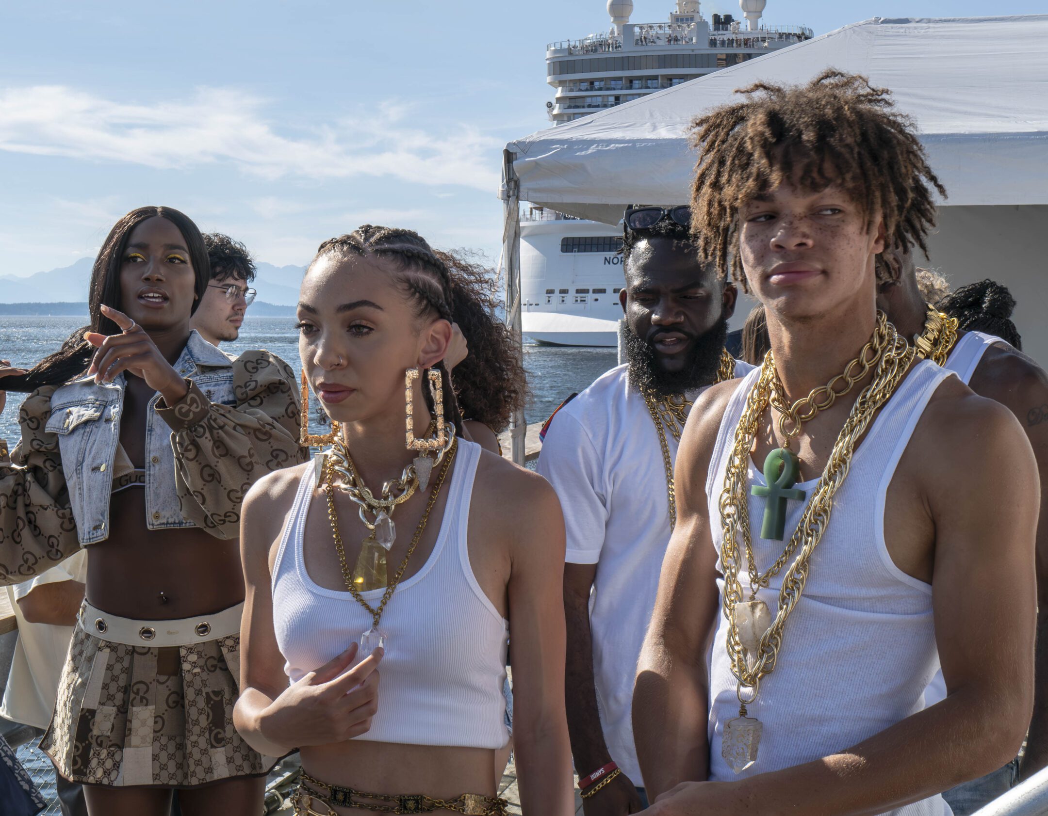 4 Black models wearing different outfits and an assortment of jewelry and gold accessories. They are outdoors at Pier 62. In the background, there is a cruise ship and the blue summer sky.
