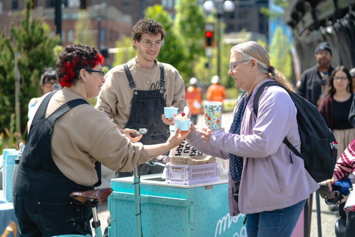 Two Molly Moon's staff members standing by their ice cream cart. One staff member is offering a sundae to a customer.
