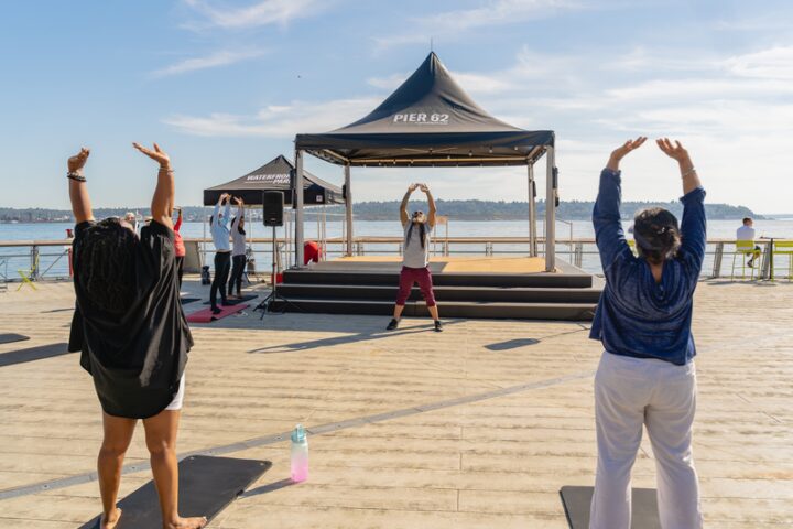 Two students stretching hands up above heads on a pier following the instruction from a teacher in the center of the photo.