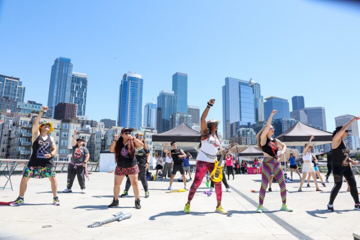 Several students participating in a fitness dance class, dressed in vibrant colors. Skyscrapers in the distance. Sun shining brightly. 