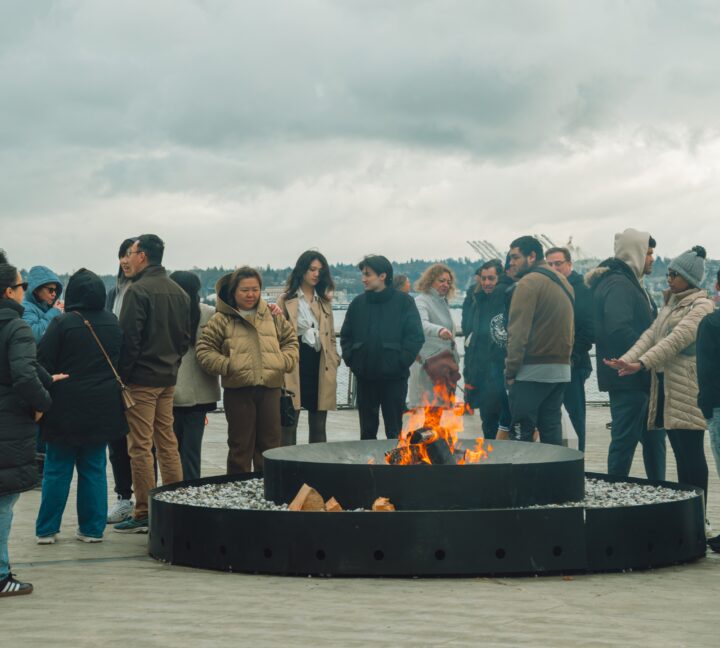 Visitors gathered around a large wood burning fire on Pier 62 during a cloudy day.