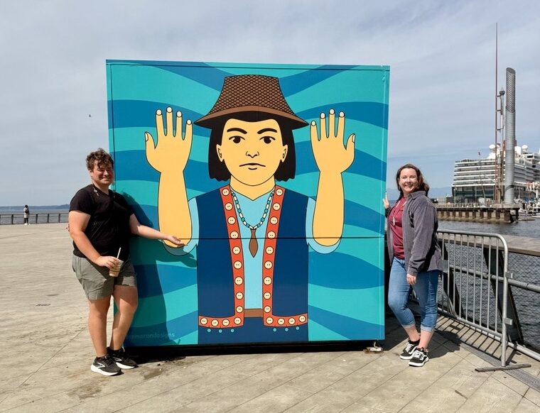 Two visitors pose on either side of a shipping container featuring a welcoming figure with raised hands, wearing traditional regalia, set against a blue wave background, part of Taylor Cameron's Currents of Connection mural on Pier 62.