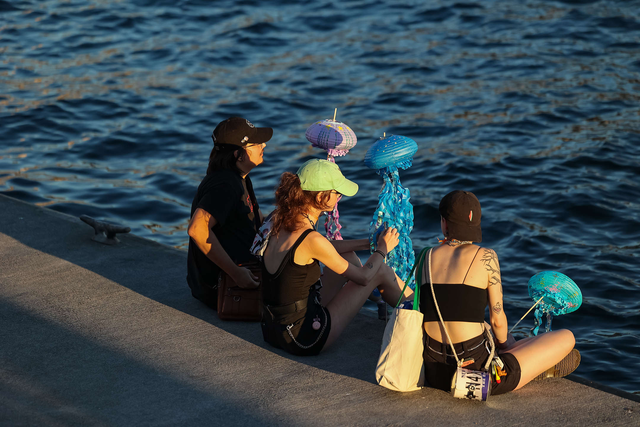 This image captures three young adults relaxing by the waterfront, each holding a colorful, handmade lantern resembling a jellyfish with long, flowing ribbons. 