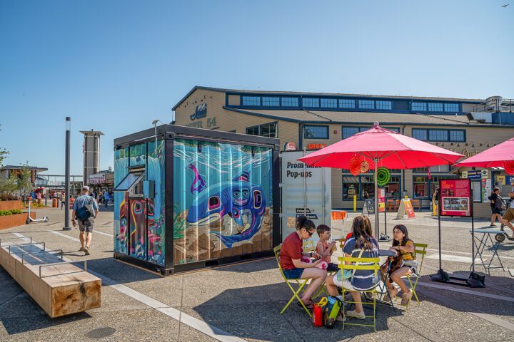 Vibrant promenade pop-up container at Waterfront Park with people sitting around a cafe table enjoying lunch.