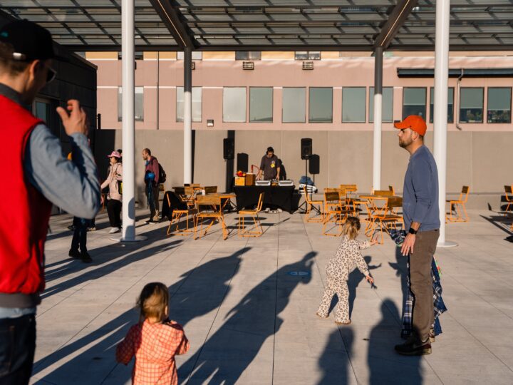 A vibrant public gathering at a covered plaza by Overlook Walk cafe with a DJ playing music, families with children dancing, and people enjoying the open space.