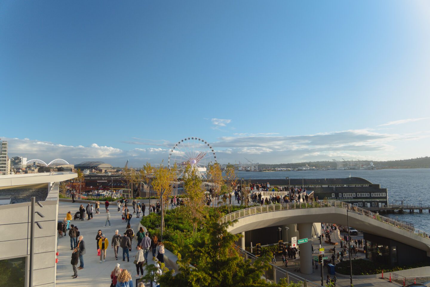 A lively urban waterfront park with people walking along a modern, landscaped pedestrian bridge. The Seattle Great Wheel and Elliott Bay are visible in the background, along with a mix of city buildings and distant stadiums.