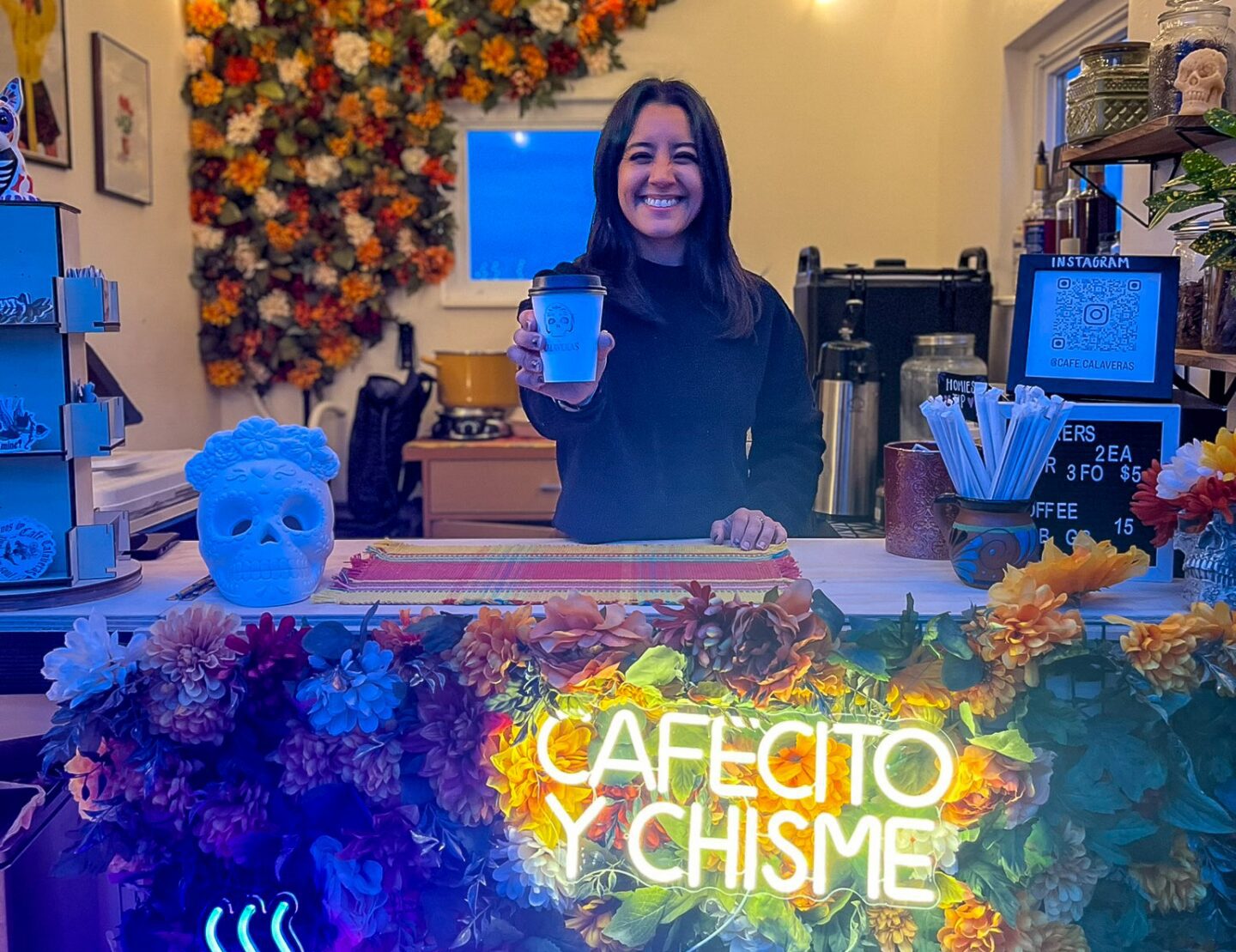 A woman, smiling, stands behind a cozy, floral-decorated café counter with a neon sign that reads “cafecito y chisme.”