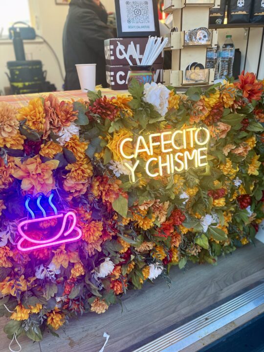 A floral stand with Café Calaveras’ recognizable neon sign that reads Cafecito y chisme. On top of the stand are various pieces of merchandise.