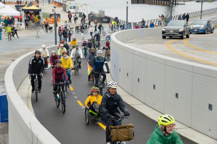 A diverse group of cyclists rides along a newly opened bike lane at Seattle’s waterfront, with people walking and gathering near vendor tents in the background. A few cars travel on the adjacent elevated roadway.