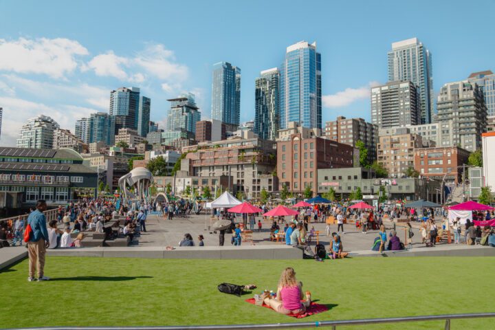 A sunny day at Waterfront Park’s Pier 58, with people lounging on the grass and strolling through a lively market filled with colorful tents. The Seattle skyline rises in the background, blending the cityscape with the waterfront.
