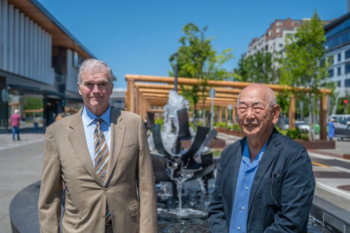 Two men stand smiling in front of the Tsutakawa Fountain at Waterfront Park on a sunny day, with the fountain and pergola structure in the background.