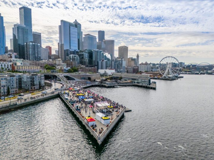 A wide aerial view of Pier 62 at Seattle’s Waterfront Park, bustling with colorful vendor tents and festivalgoers. The Seattle skyline rises in the background, with the Seattle Great Wheel and Elliott Bay visible under a partly cloudy sky.