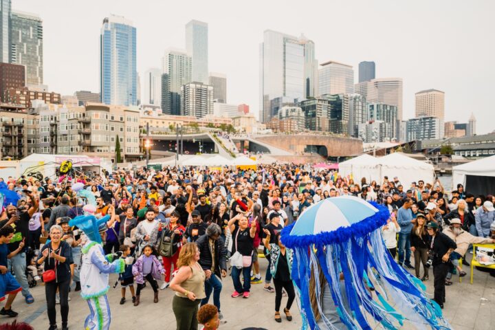 A large, energetic crowd gathers on Pier 62 during a waterfront celebration in Seattle. People of all ages are dancing, smiling, and moving together.