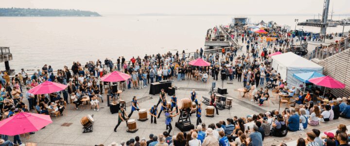 A large crowd gathers at the Salish Steps at Overlook Walk, watching a group of drummers perform at the center. People fill the stepped seating, upper walkways, and open plaza space, with bright pink umbrellas and small tables scattered throughout.
