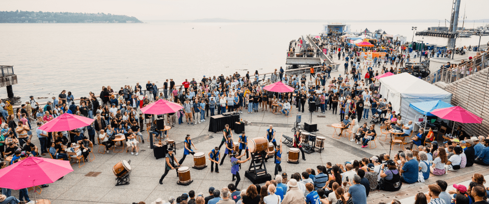 A large crowd gathers at the Salish Steps at Overlook Walk, watching a group of drummers perform at the center. People fill the stepped seating, upper walkways, and open plaza space, with bright pink umbrellas and small tables scattered throughout.