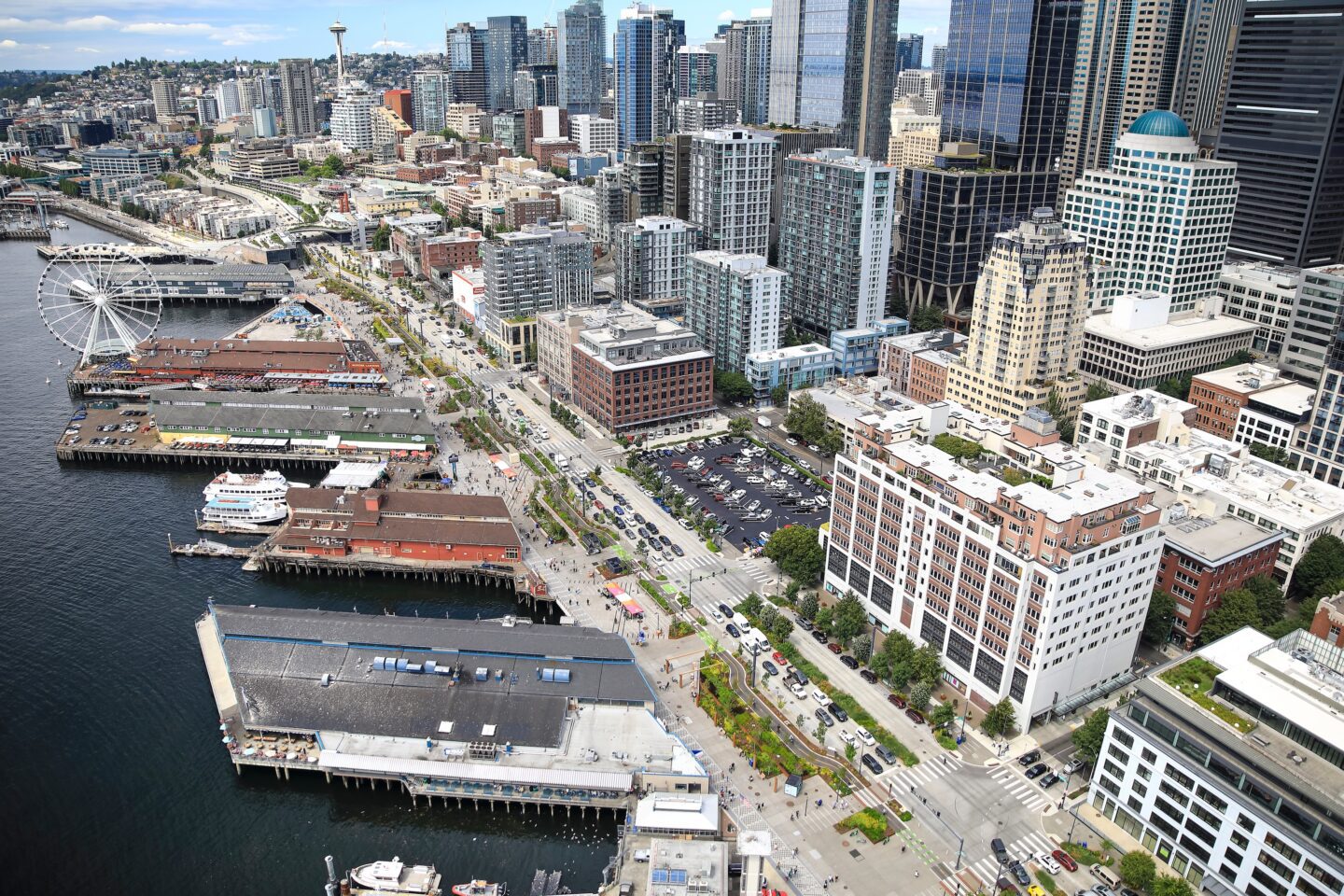 Aerial view of Seattle’s central waterfront and downtown skyline, showing piers, a waterfront promenade, ferries, busy streets, and tall buildings stretching inland on a clear day.