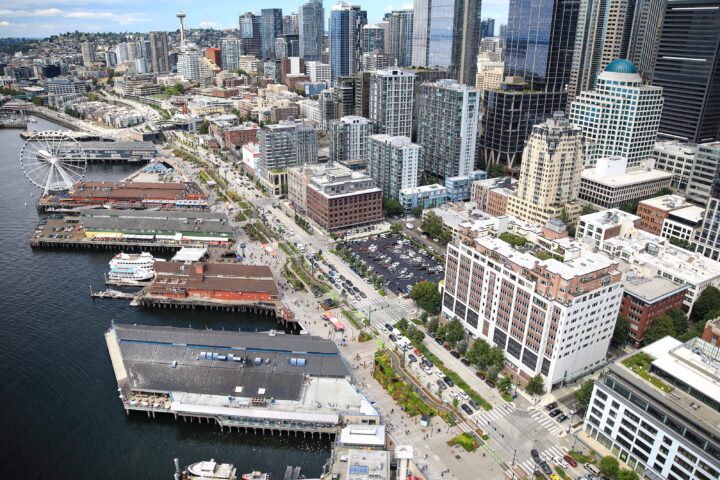 Aerial view of Seattle’s central waterfront and downtown skyline, showing piers, a waterfront promenade, ferries, busy streets, and tall buildings stretching inland on a clear day.