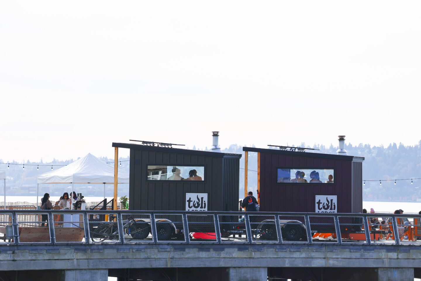 Two Tuli Lodge sauna cabins sit side by side on Pier 62, with guests visible through the windows and the Salish Sea and tree-lined shoreline in the background.