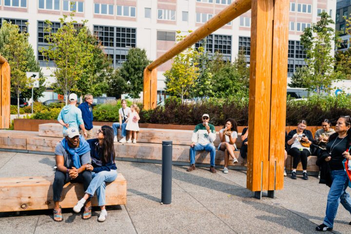 A diverse group of visitors sit and gather on wooden benches near the Tuazon log house installation at Waterfront Park on a sunny day.