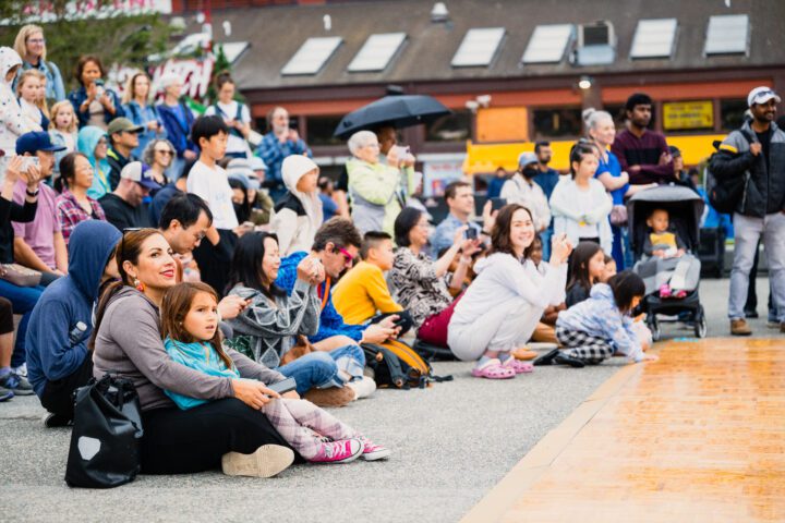 A diverse crowd of families, children and community members gather at Waterfront Park's Grand Opening Celebration, many seated on the ground watching a performance.