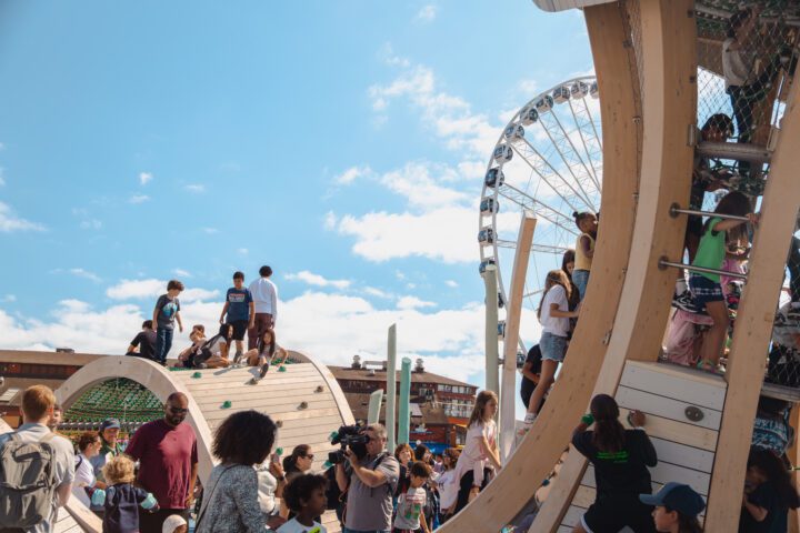 Children and families climb and play on the Jellyfish Playground at Pier 58 with the Seattle Great Wheel visible in the background under a bright blue sky.