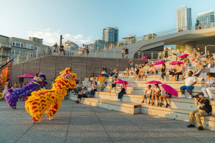 A vibrant lion dance performance takes place at the Salish Steps amphitheater at Waterfront Park as a crowd watches from the steps under the Seattle skyline.