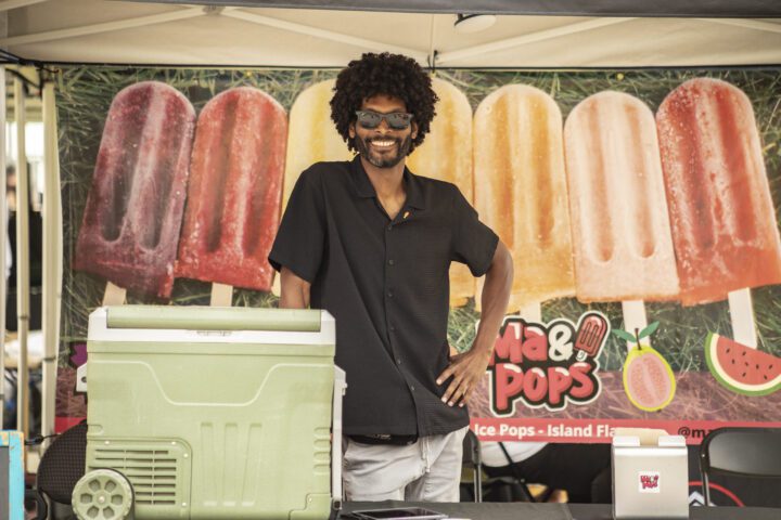 A smiling vendor stands in front of the Ma & Pops ice pop booth with a green cooler.